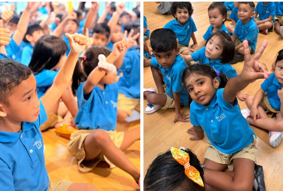 Children in blue polo shirts sit on a wooden floor, many raising hands; one girl flashes a peace sign.